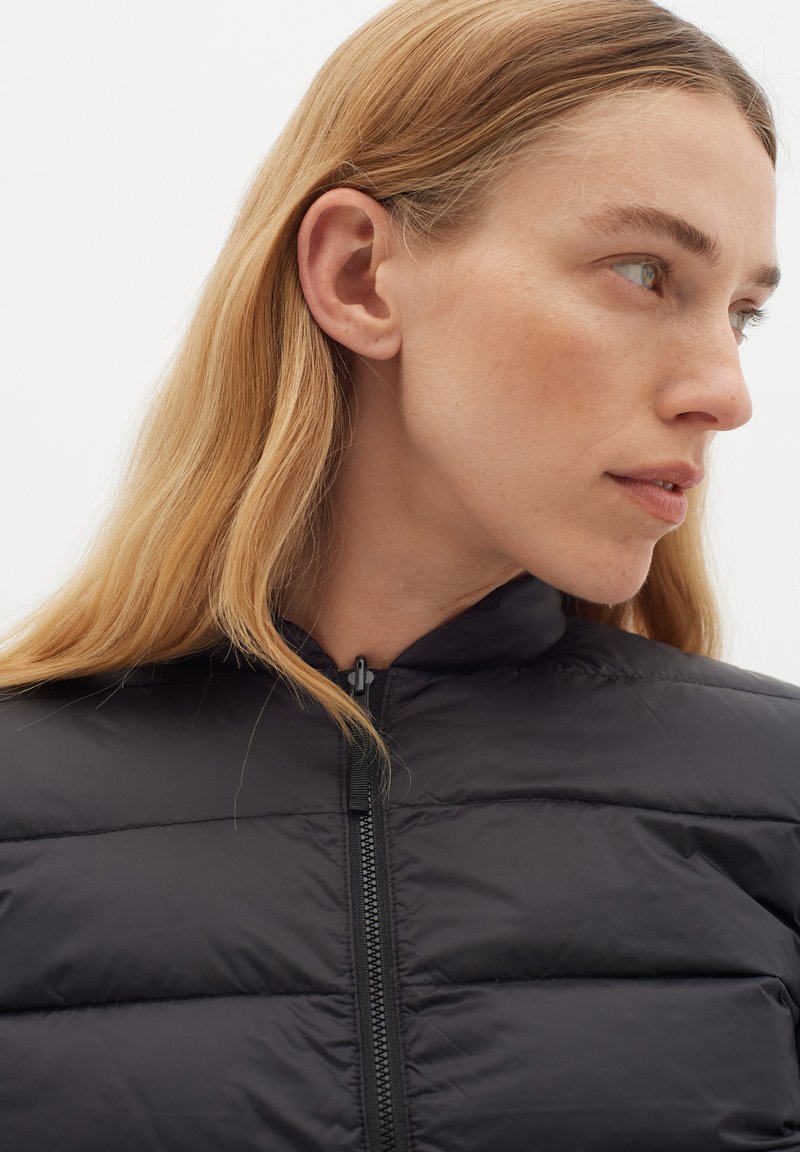 Schwarze Steppjacke mit gestepptem Muster, hohem Kragen und Frontreißverschluss. Der Stoff wirkt leicht und glatt. Das Modell hat weiche, fließende Haare.