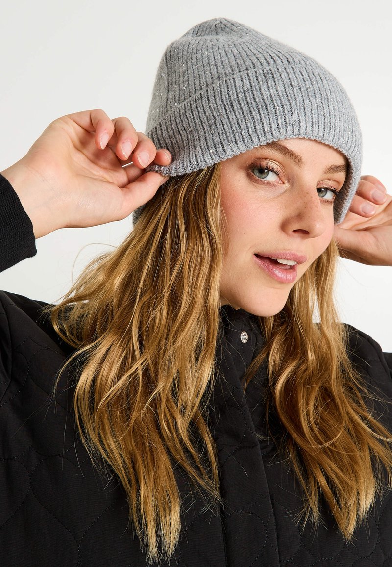 Gorro de punto acanalado gris con una textura suave, que presenta sutiles destellos. La modelo ajusta el gorro, vistiendo una chaqueta acolchada negra.