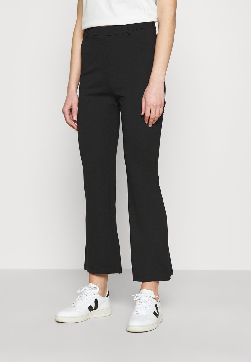Personne portant un pantalon noir droit et des baskets blanches avec un logo « V » noir, debout devant un fond clair uni.