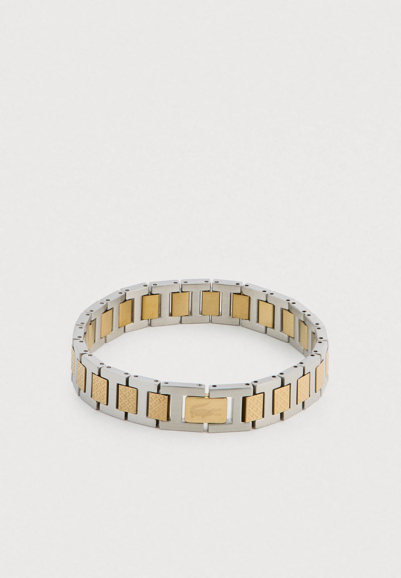 Metāla aproce ar mainīgām sudraba un zelta sekcijām. Teksturētie zelta akcenti ar gludu aizdari. Gluda, taisnstūrveida dizains.