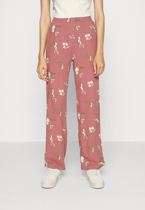 Mujer con pantalones rosas de talle alto y pierna ancha, con estampado floral blanco y deportivas blancas, de pie frente a un fondo blanco liso.