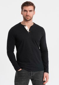 Svart långärmad skjorta med rund halsringning, med vit knäppning och tre bruna knappar. Bomullstyg med en jämn textur.