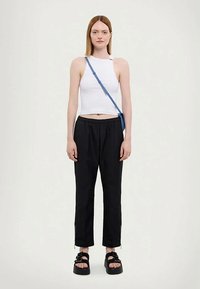 Weißes, kurz geschnittenes Tanktop, schwarze tapered Hose mit elastischem Bund und Reißverschlüssen und schwarze Sandalen mit breiten Riemen. Blauer Umhängetasche.