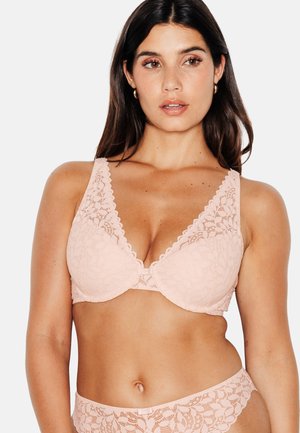 Femme portant un soutien-gorge en dentelle rose clair et des sous-vêtements assortis, montrant le haut du corps et une expression faciale neutre sur un fond uni.