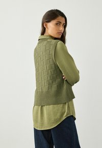 Grön stickad väst med en strukturerad, rutmönstrad design. Bärs över en matchande olivgrön skjorta och mörkblå jeans. Ribbad nederkant och ärmlös design.