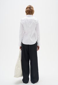 Henkilö, jolla on aaltoilevat vaaleanruskeat hiukset, seisoo selinpäin ja pukeutunut valkoiseen pitkähihnaiseen paitaan, mustiin leveälahkeisiin housuihin ja pitää kädessään suurta valkoista kangaskassia.