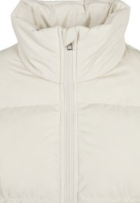 Lyse beige pufferjakke med høj krave, frontlynlås, quiltet tekstur og minimale syninger.