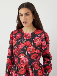 Schwarzes Blumenkleid mit roten und rosa Mohnblumenmustern, strukturiertem Stoff, langen Ärmeln und gerafftem Taillenbereich. Goldene Hoop-Ohringe runden den Look ab.