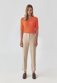 Orange short-sleeve knit sweater with a ribbed texture paired with beige tailored trousers and brown slip-on shoes, standing against a light background.