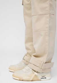 Pantalon cargo beige à coupe décontractée et ourlet retroussé, associé à des baskets de couleur claire dotées d'une tige translucide et du détail "AIR" sur le talon.
