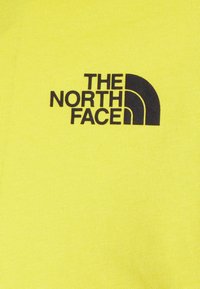 Żółta tkanina z czarnym logo z zakrzywionymi elementami z napisem „THE NORTH FACE”. Tekstura wygląda na gładką i dzianą.