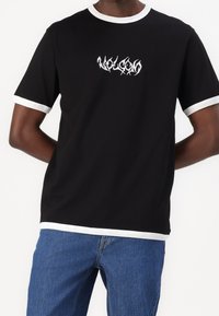 Zwarte T-shirt met witte geribbelde kraag en mouwranden, met een gestileerd wit logo op de borst. Zacht materiaal, casual pasvorm.