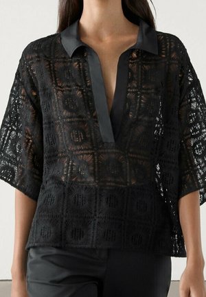 Femme portant un haut en dentelle noire avec des motifs géométriques et un col en satin, associé à un pantalon noir, sur un fond uni.