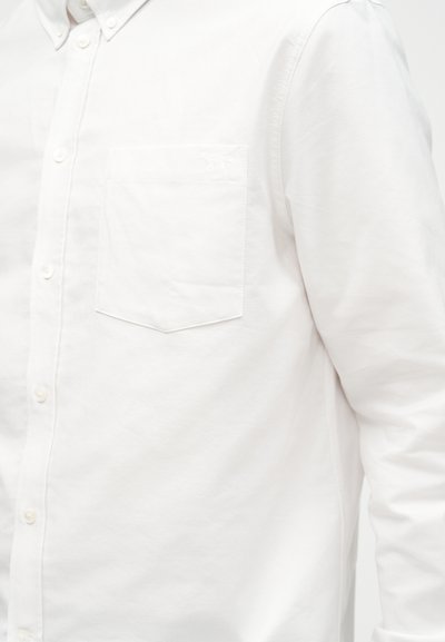 Άνδρας που φοράει λευκό πουκάμισο με μακριά μανίκια και κουμπιά, με τσέπη στο στήθος, εμφανιζόμενος από το στήθος μέχρι το μέσο του βραχίονα πάνω σε απλό φόντο.