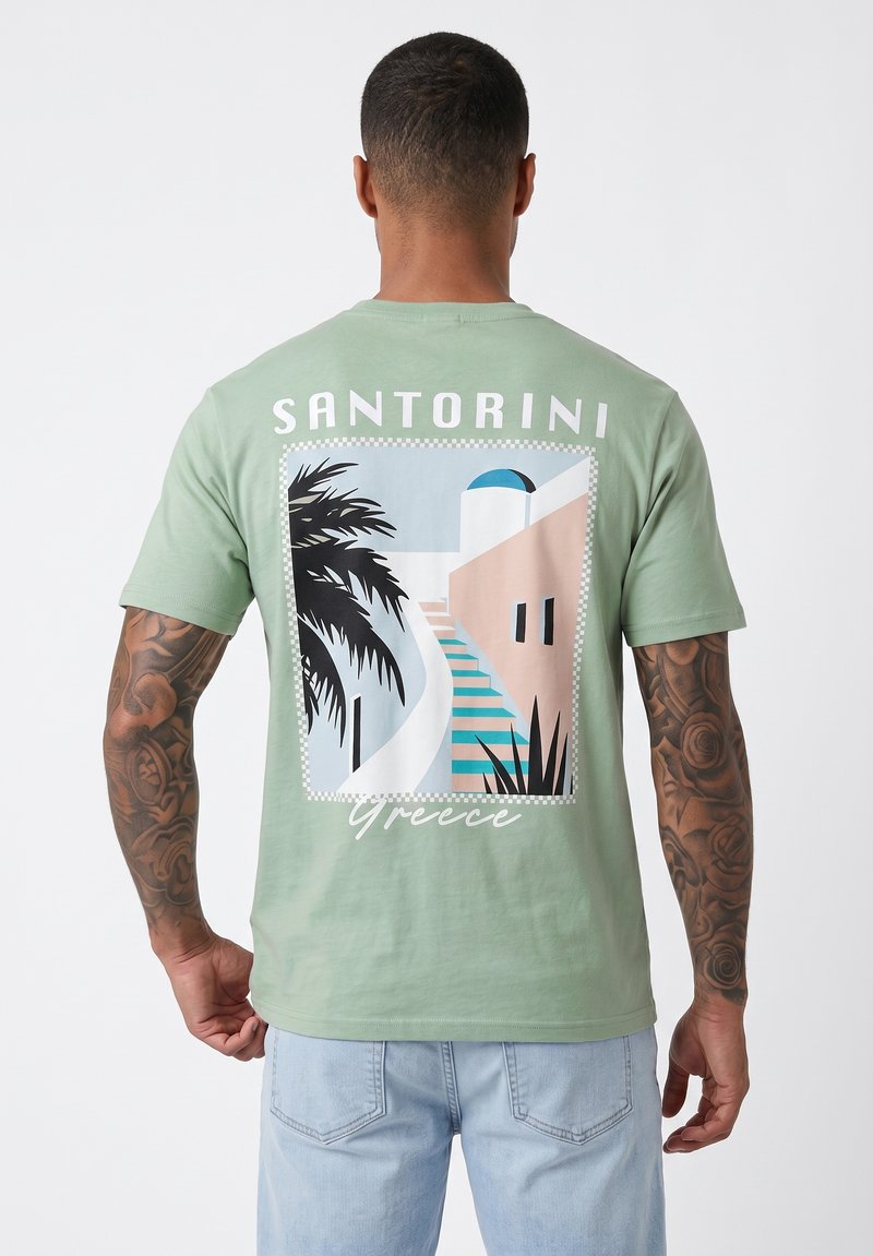 Homme aux bras tatoués portant un t-shirt vert avec un graphisme de Santorin, Grèce, montrant des escaliers, un palmier et des bâtiments blancs avec un dôme bleu.