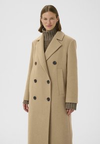 Cappotto beige doppiopetto in misto lana, con bottoni neri, ampie rever e tasche laterali. Il modello indossa un maglione a coste sotto.