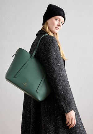 Femme portant un bonnet en tricot noir et un manteau texturé foncé, portant un grand sac cabas vert sur l'épaule sur fond gris uni.