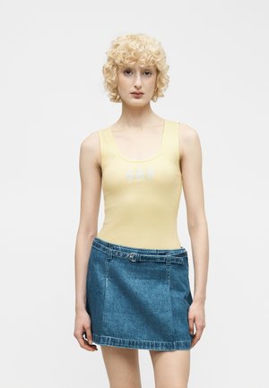 Młoda kobieta z krótkimi kręconymi blond włosami, ubrana w żółtą, bezrękawnik i niebieską dżinsową spódnicę z paskiem na tle gładkiego tła.