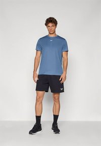 Jonge man staat en draagt een blauw sportshirt, zwarte korte broek, zwarte sokken en zwarte sneakers tegen een effen achtergrond.