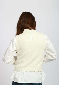 Gilet lavorato a maglia colore crema con collo alto, orlo a coste e dettagli a chiave sul retro, indossato sopra una camicia bianca con maniche lunghe.