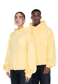 Sudaderas amarillas de tela suave con un bolsillo frontal y el logo bordado "MULTIPLY". Los modelos llevan jeans oscuros, mostrando un corte relajado.