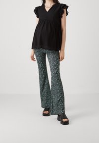 Top nero texturizzato con maniche arricciate e scollatura a V, abbinato a pantaloni verdi svasati con piccoli motivi floreali. Sandali con tacco nero.