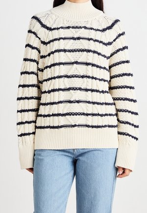 Roomkleurige kabelgebreide trui met horizontale marineblauwe strepen, gedragen met lichtblauwe spijkerbroek, waarbij de torso en armen zichtbaar zijn.