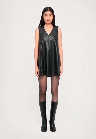 Vrouw in een mouwloze zwarte leren mini-jurk, doorzichtige zwarte panty’s en zwarte leren laarzen tot aan de knie, staand tegen een effen achtergrond.