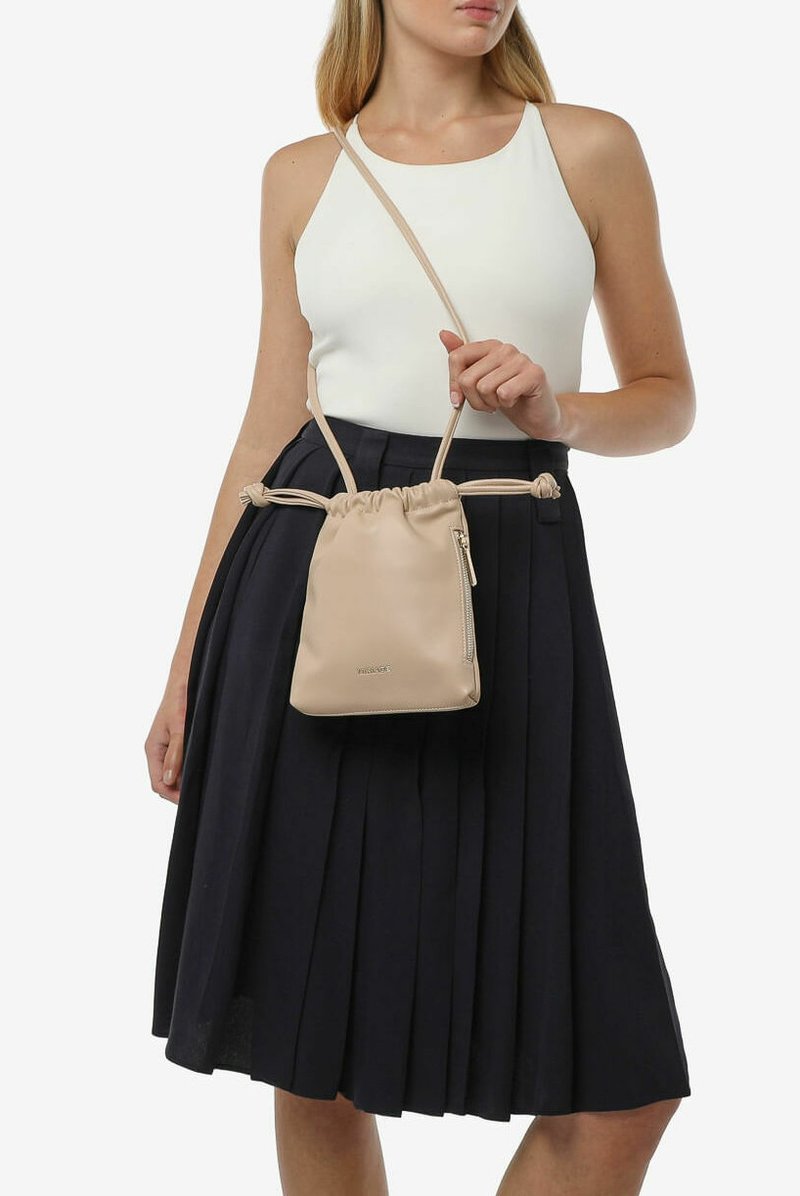 Bolso de piel beige con cordón y un bolsillo lateral con cremallera, textura suave y correas ajustables, sostenido por una persona que lleva una blusa blanca y una falda oscura.