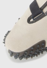 Beige suede shoe with a textured gray rubber sole featuring sharp, tapered spikes. Black strap across the upper adds contrast and stability.