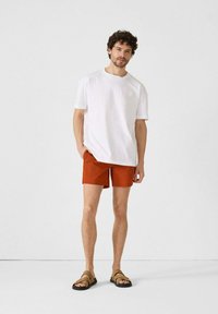 T-shirt bianco a maniche corte, pantaloni shorts arancioni con un motivo testurizzato, sandali neri con cinturini marroni, in piedi contro uno sfondo neutro.