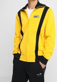 Gul zip-up jacka med svarta ränder och krage. Logotyp på övre vänstra bröstet. Mjuk material, avslappnad passform, ihop med svarta byxor.