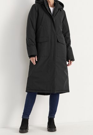 Personne portant un long manteau d'hiver noir à capuche, un jean bleu et des bottines noires, debout devant un fond blanc.