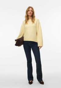 Pull-over jaune à coupe décontractée, ourlet côtelé et épaules tombantes, associé à un jean évasé bleu foncé. Tenant une pochette marron.