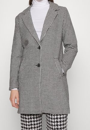 Femme portant un blazer pied-de-poule noir et blanc avec deux boutons noirs et un pantalon assorti, un col roulé blanc en dessous.
