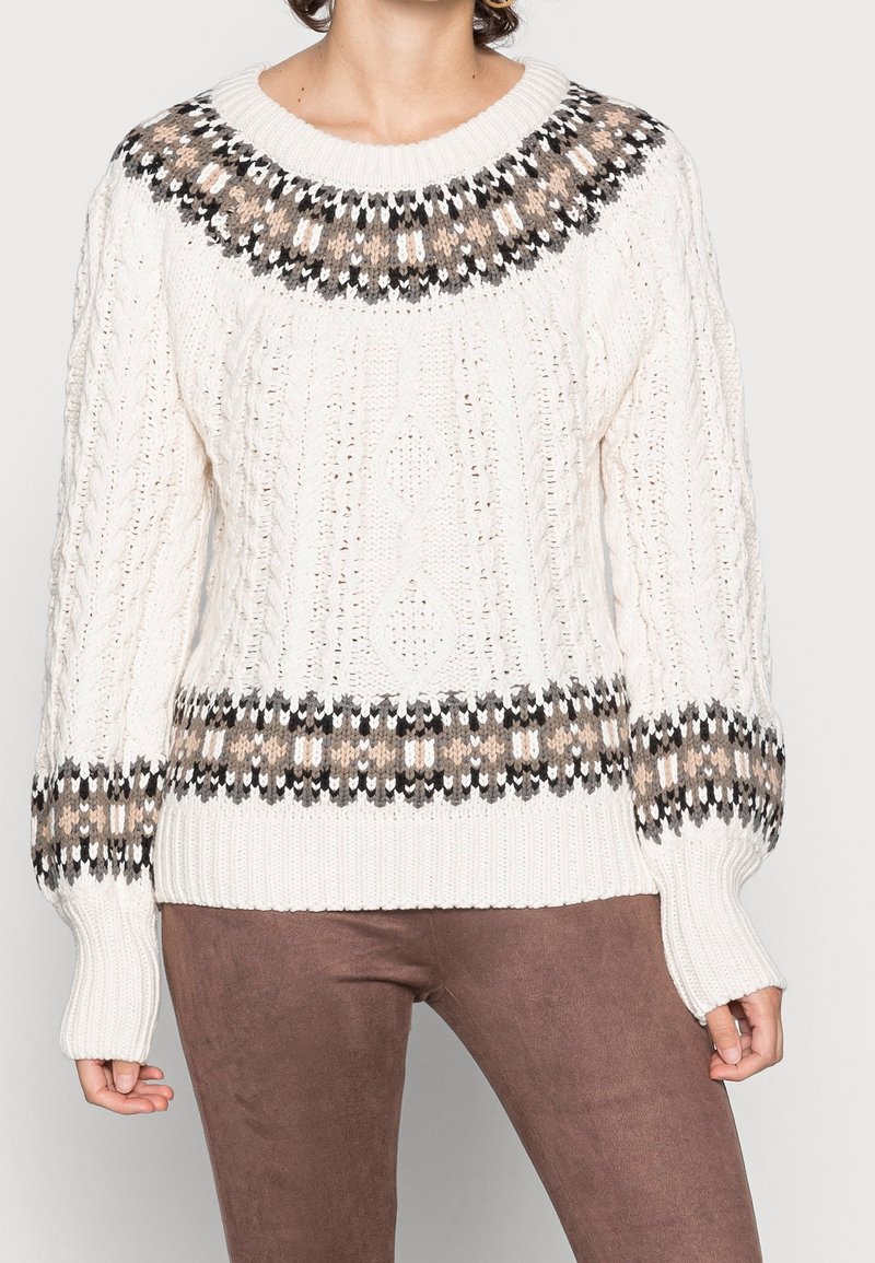 Femme portant un pull en maille blanc avec un empiècement, des poignets et un ourlet à motifs marron, beige et noir, associé à un pantalon marron.