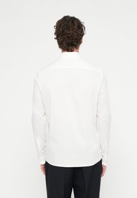 Λευκό πουκάμισο μακρυμάνικο από λείο ύφασμα, με κλασικό γιακά και κυρτή ραφή στο κάτω μέρος. Η πίσω όψη δείχνει λεπτομέρειες με κουμπιά στα μανίκια.