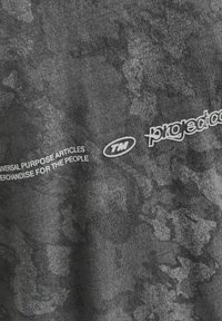 Hall hõbehallist puuvillase t-särgist, millel on tekstuuriline ja kirju disain, sisaldab valget teksti, mis ütleb "UNIVERSAALSUSE EESMÄRGILISTE ARTIKLITE" ja "projekt".