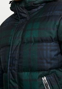 Puffjacka med en mörkgrön och marinblå rutigt mönster. Tillverkad av ett texturerat tyg har den en hög krage och en ficka med dragkedja.