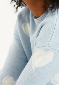 Jasnoniebieski sweter wykonany z miękkiego materiału, z fakturowanymi białymi wzorami serc. Zbliżenie pokazuje szczegóły ściągacza kołnierza i rękawów.