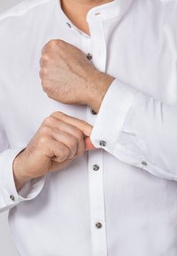 Chemise blanche à boutons avec un col mao et des boutons gris. Le tissu semble lisse, et les poignets sont en train d'être boutonnés.
