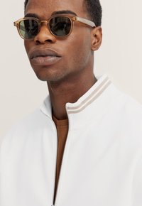 Lightweight, clear sunglasses with green lenses. The wearer dons a white jacket with brown accents and a brown shirt underneath.