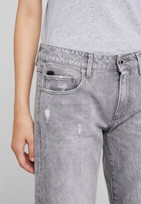 Jeans de denim gris con un ajuste relajado, que presentan detalles desgastados, un cierre de botón y un pequeño bolsillo cerca de la cintura. Tejido texturizado.