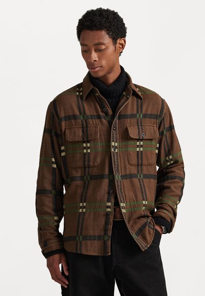 Camisa a cuadros marrón con líneas verdes y negras, dos bolsillos en el pecho y diseño de botones. Llevada sobre un suéter de cuello alto negro. Textura suave.