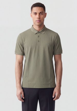 Uomo che indossa una polo verde oliva a maniche corte con tre bottoni e pantaloni scuri, in piedi davanti a uno sfondo chiaro uniforme.