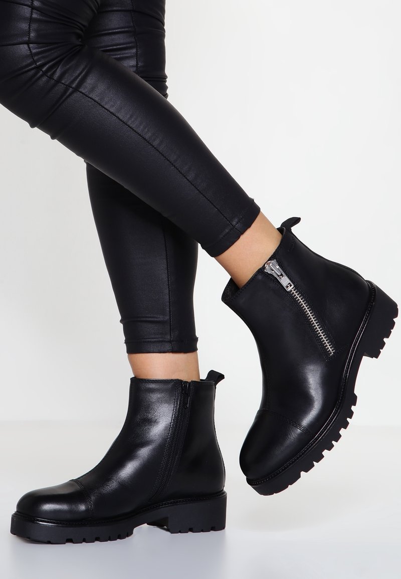 Bottines en cuir noir avec une semelle épaisse, fermeture éclair sur le côté et languette de tirage. Surface lisse avec un léger éclat, semelle extérieure texturée contrastante.