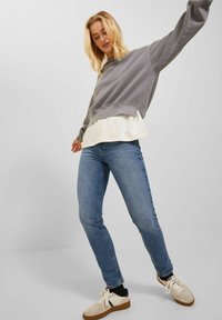 Grauer Pullover über einem weißen Oberteil, kombiniert mit blauen Skinny-Jeans und beigen Sneakers. Schlichtes Design, lockere Passform und minimale Akzente.