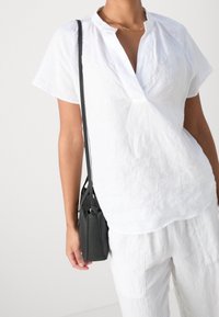 Chemise blanche en lin à manches courtes et col en V, portée avec un pantalon blanc ample. Sac bandoulière noir avec une lanière d'épaule et une texture subtile.