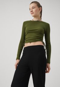 Long-sleeve olive green top with a ribbed texture, featuring side drawstrings. Paired with black ribbed wide-leg pants.