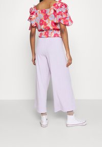 Bluse mit Blumenmuster und Rüschensleeves, kombiniert mit helllila weit geschnittenen Hosen und weißen Turnschuhen, von hinten gezeigt.